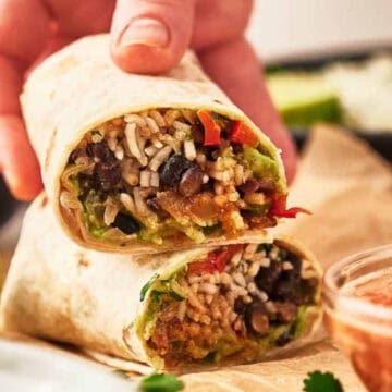 A hand holds a cut vegan burrito filled with rice, beans, and veggies, near salsa and lime wedges.