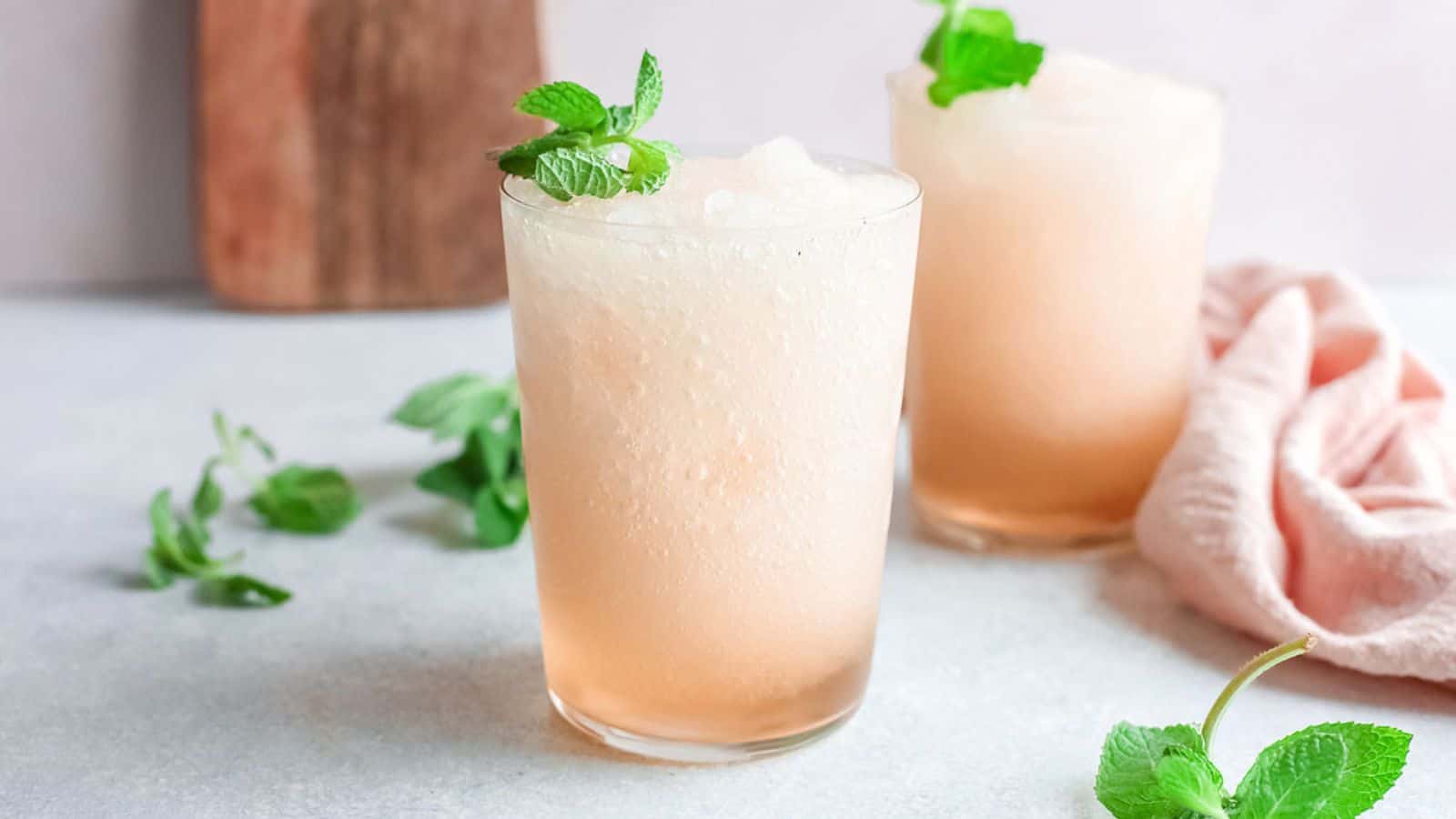 Two frosty peach-colored drinks in glasses, garnished with mint leaves, on a light surface with mint sprigs nearby.