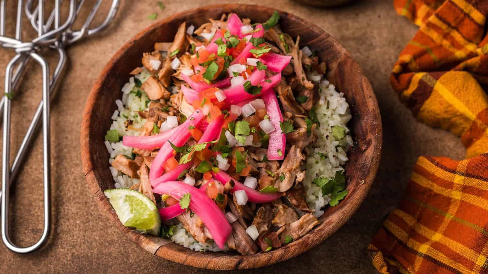 A wooden bowl of rice topped with shredded meat, pickled onions, salsa, cilantro, and a lime wedge.
