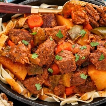 Beef stew with potatoes, carrots, and herbs served over noodles in a black dish, with a spoon and garnish.
