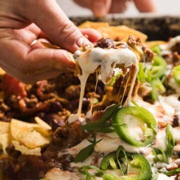A hand lifting cheesy nachos topped with jalapeños, beans, ground meat, and sour cream from a tray—perfect cinco de mayo appetizers for your celebration.