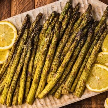 Grilled asparagus spears on a platter with lemon slices, set on a wooden table next to a yellow napkin.