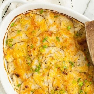 Oval dish of cheesy scalloped potatoes with herbs, inspired by vintage recipes, sits beside a wooden spatula on a striped towel.