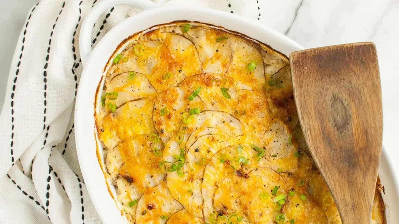 Oval dish of cheesy scalloped potatoes with herbs, inspired by vintage recipes, sits beside a wooden spatula on a striped towel.