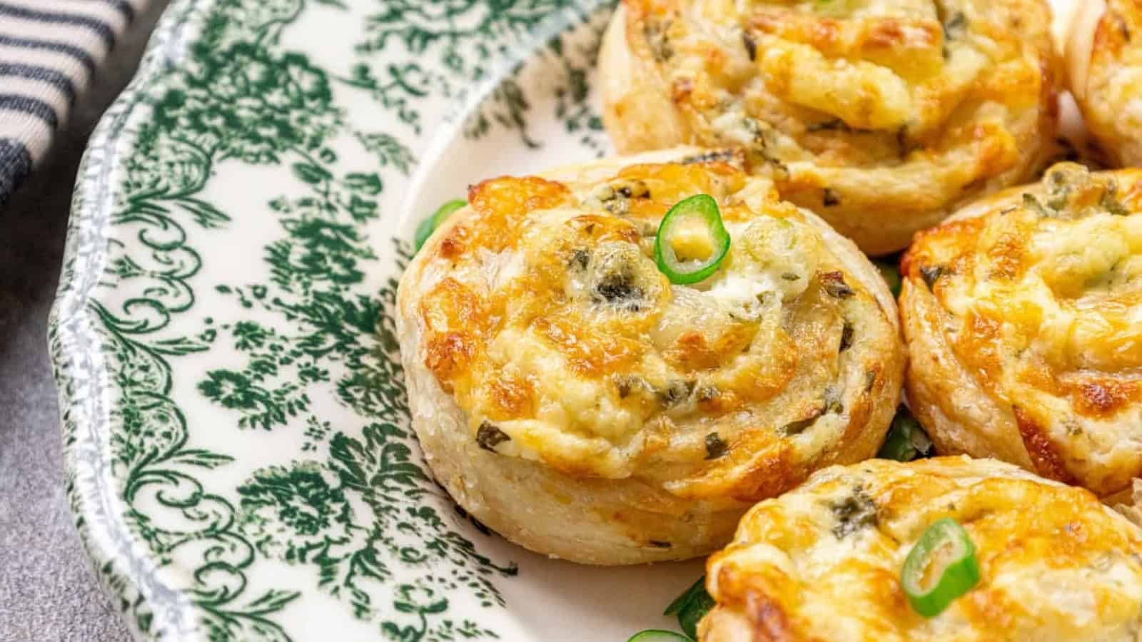 Golden, cheesy puff pastry pinwheels with herbs on a decorative green and white plate.
