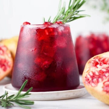 Glass of pomegranate drink with ice, garnished with rosemary, surrounded by fresh pomegranate halves.