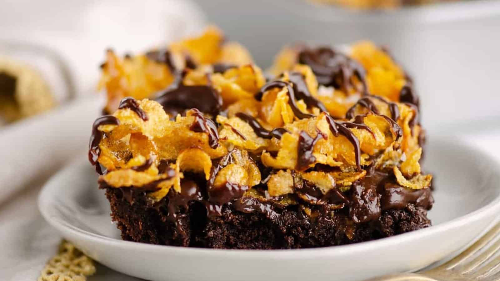 A brownie topped with crunchy cornflakes and drizzled chocolate on a white plate.