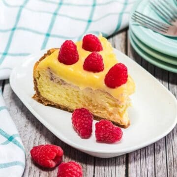 Slice of cheesecake topped with raspberries on a white plate, beside stacked plates, forks, and a checkered cloth.