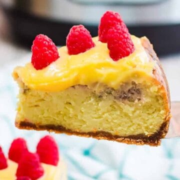 A slice of cheesecake topped with yellow cream and fresh raspberries held up on a spatula.