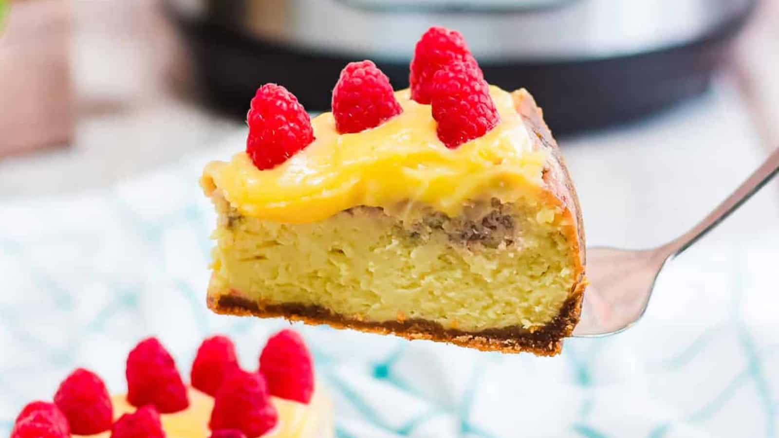 A slice of cheesecake topped with yellow cream and fresh raspberries held up on a spatula.