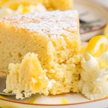 A slice of lemon cake with powdered sugar, whipped cream, and a lemon slice on a plate.