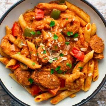 A bowl of penne pasta with sausage, chicken, bell peppers, and tomato sauce, garnished with parsley and cheese.