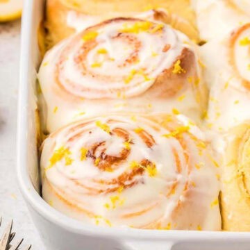 Iced lemon sweet rolls in a white baking dish, topped with lemon zest, with a lemon reamer nearby.
