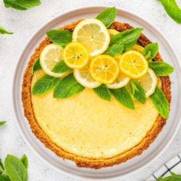 Lemon cheesecake topped with lemon slices and mint leaves on a plate, surrounded by lemons and mint.
