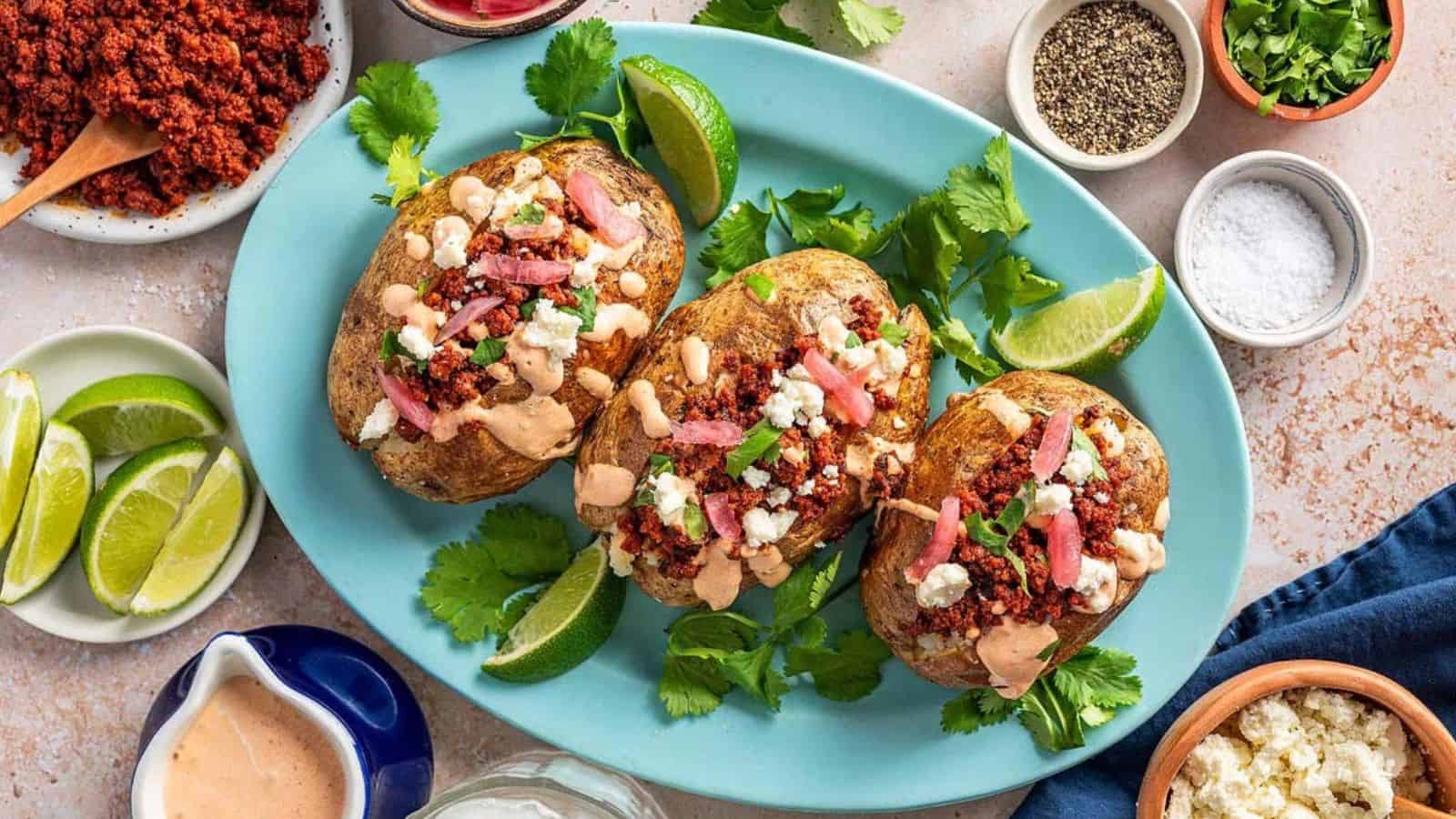 Three loaded baked potatoes topped with chorizo, sauce, cheese, pickled onions, and lime wedges on a blue platter.