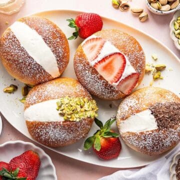 Round cream-filled pastries topped with strawberries, pistachios, and chocolate, on a plate with coffee and ingredients.