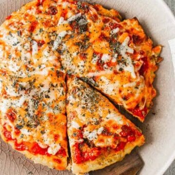 A round cheesy pizza with herbs, one slice slightly pulled out on a white plate on a wooden table.