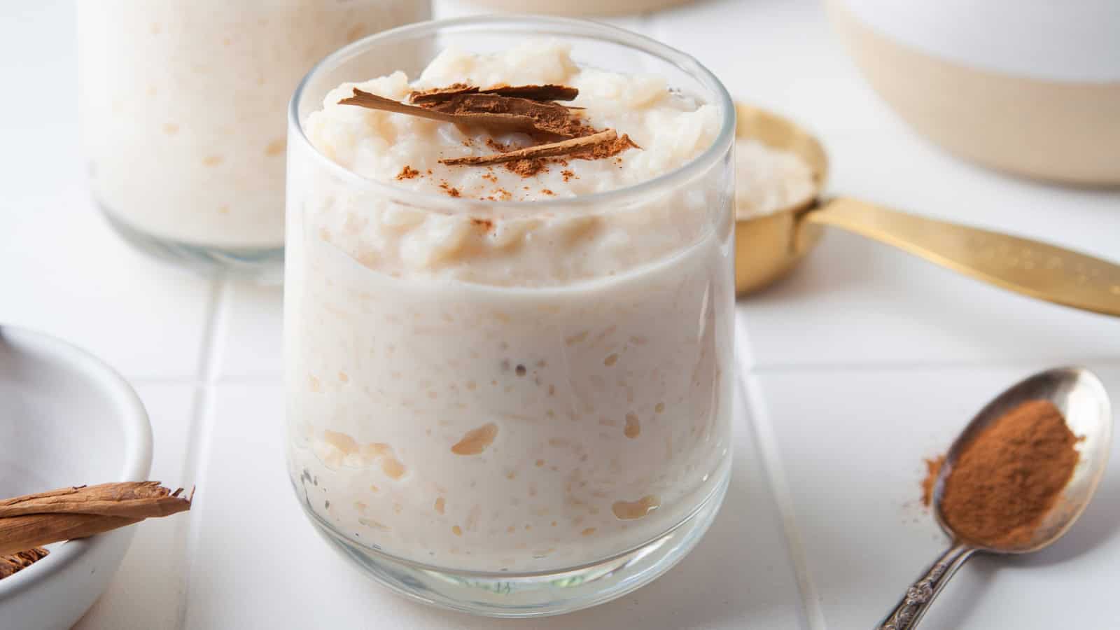 A glass of rice pudding topped with cinnamon sticks and ground cinnamon on a white table.