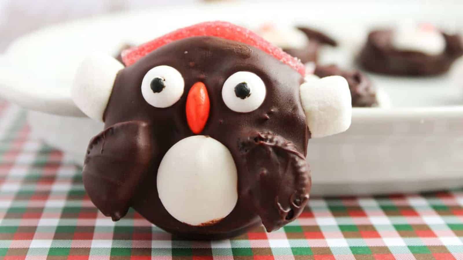 A chocolate-covered treat shaped like a penguin, with candy eyes, a red beak, and white marshmallow wings, on a checkered tablecloth.