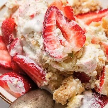 A plate of strawberry shortcake topped with whipped cream and fresh strawberry slices, with a spoon beside it.