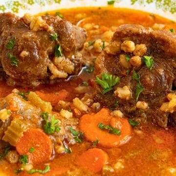 A bowl of oxtail soup with carrots, celery, rice, and herbs in a rich, red broth on a patterned plate.