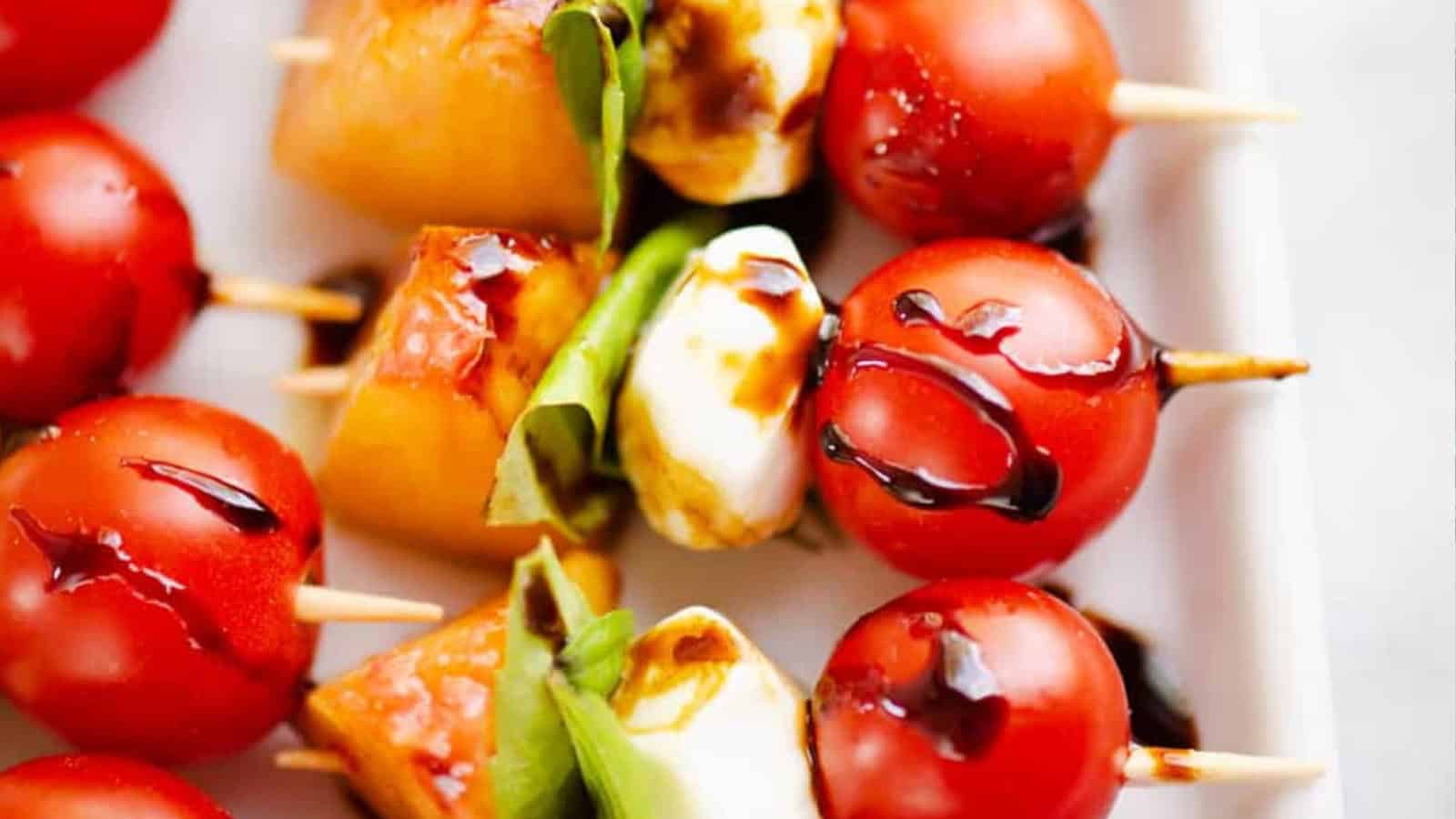Skewers with cherry tomatoes, mozzarella, basil, and melon, drizzled with balsamic glaze on a white plate.