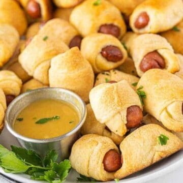 Mini pigs in a blanket, a classic among retro appetizers, are served on a platter with dipping sauce and garnished with fresh herbs.