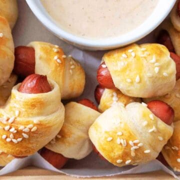 These retro appetizers feature mini pigs in a blanket with sesame seeds on top, served alongside a flavorful bowl of dipping sauce.
