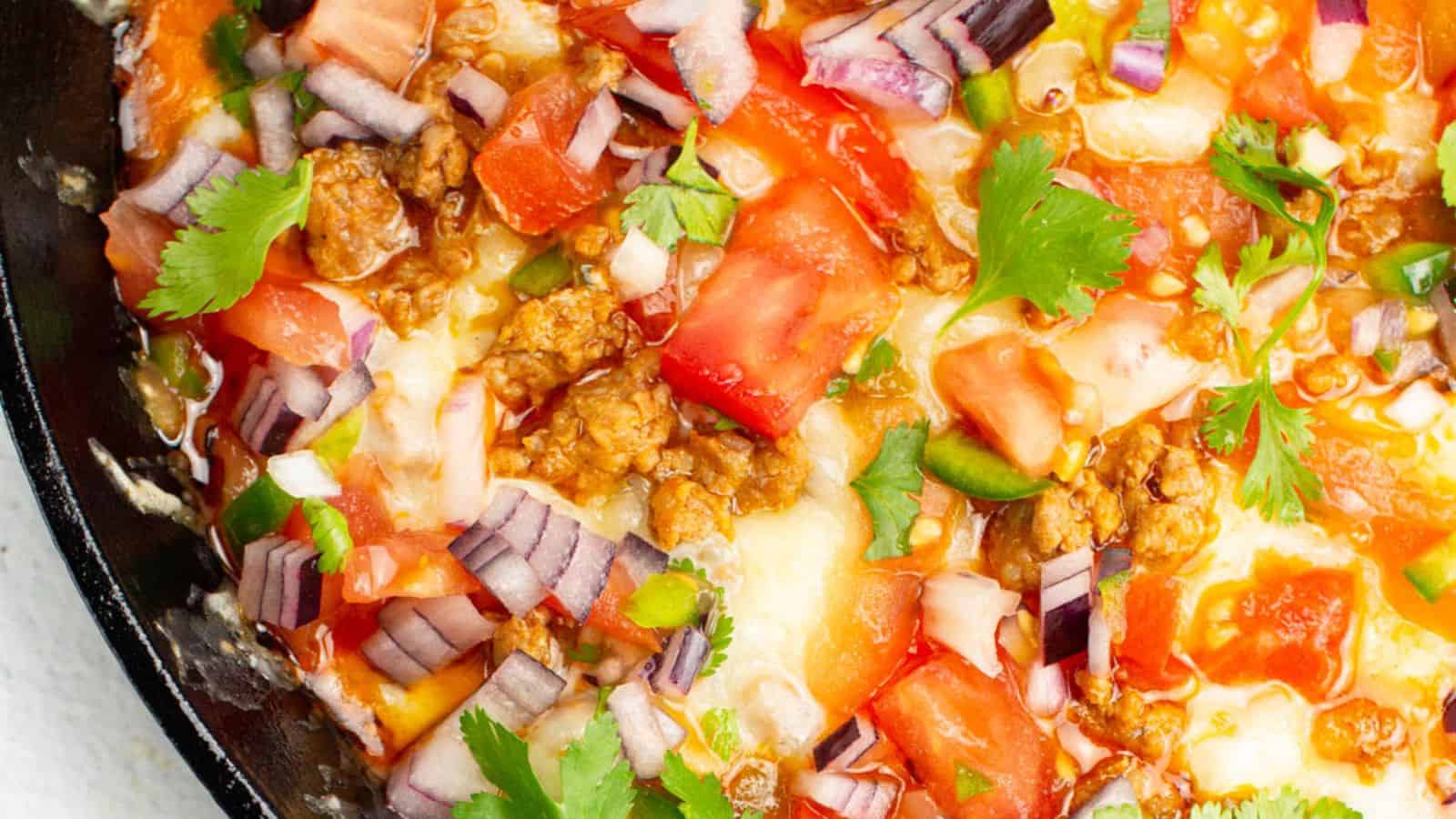 Close-up of a cheesy skillet dip topped with ground meat, diced tomatoes, red onions, jalapeños, and cilantro.