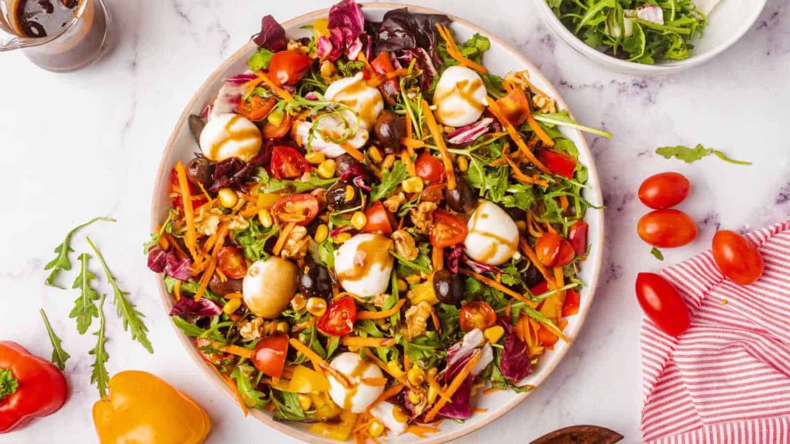 A rainbow salad with lettuce, tomatoes, carrots, olives, corn, and mozzarella balls is drizzled with dressing.