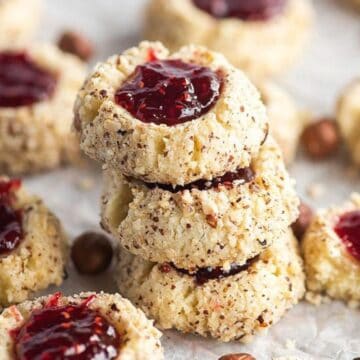 A stack of thumbprint cookies filled with red jam, surrounded by more cookies on parchment paper.
