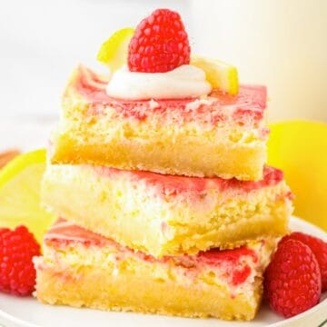 Three lemon raspberry dessert bars stacked on a plate, topped with cream, raspberries, and lemon slices.