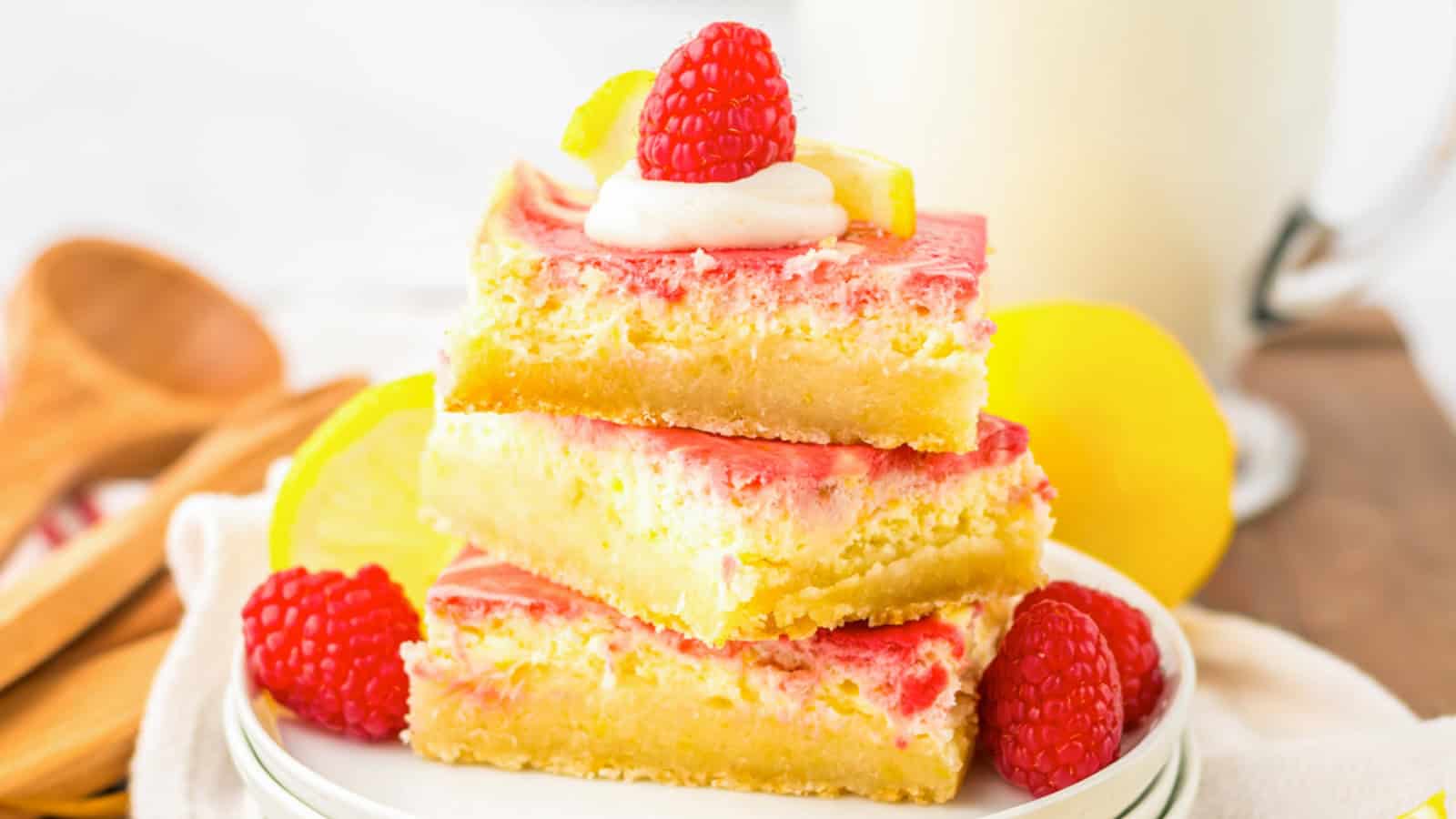 Three square lemon raspberry bars stacked elegantly on a plate. Each dessert features a yellow lemon layer topped with a pink raspberry layer. A dollop of cream adorned with a raspberry and lemon slice crowns the top, surrounded by extra raspberries and slices nearby.