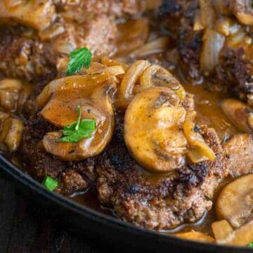 Ground beef patties topped with sautéed mushrooms and onions in a skillet with brown gravy.