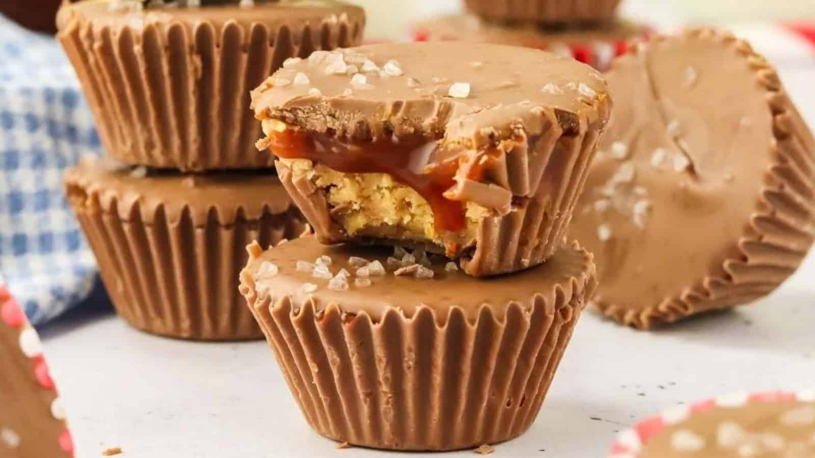 Chocolate cups with a peanut butter and caramel filling, topped with sea salt, are stacked on a table.