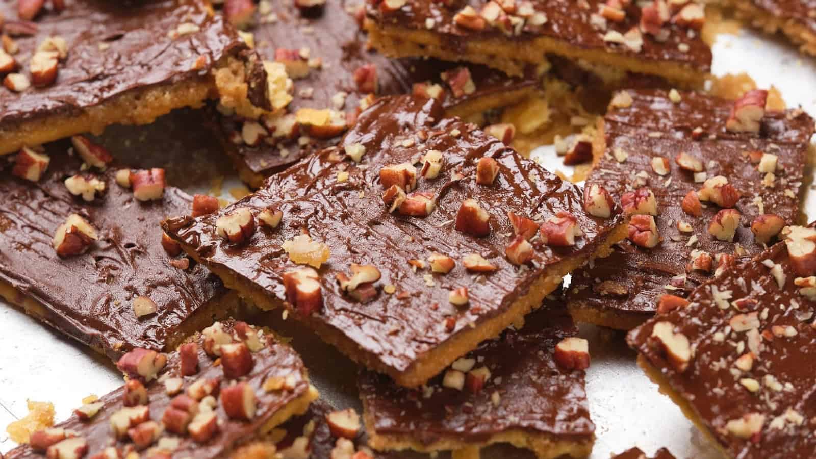 Close-up of saltine toffee squares topped with chocolate and chopped nuts on a silver surface.