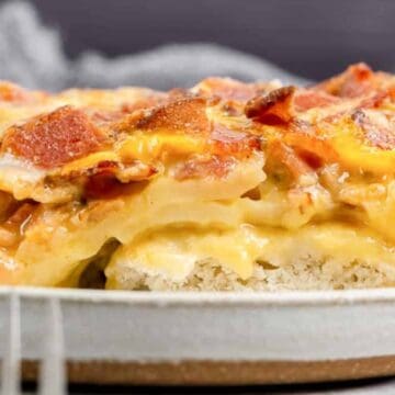A close-up of a cheesy casserole slice with bacon on top, served on a plate with a fork nearby.