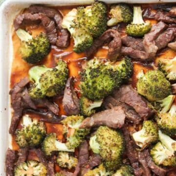 Sheet pan with cooked beef strips, broccoli florets, and sauce, next to a bowl of sesame seeds.