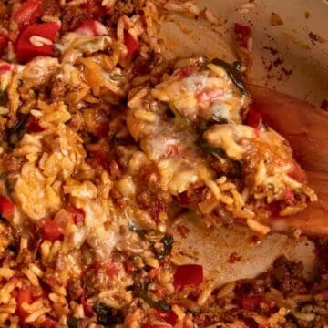 A wooden spoon scoops cheesy rice with ground meat and red bell peppers from a casserole dish.