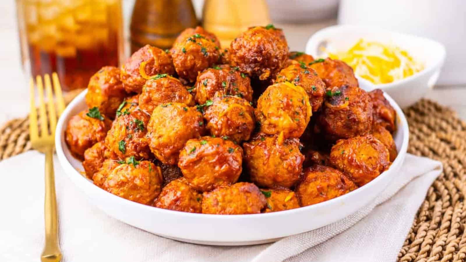 A bowl of saucy meatballs garnished with herbs, with a fork and a small bowl of shredded cheese beside it.