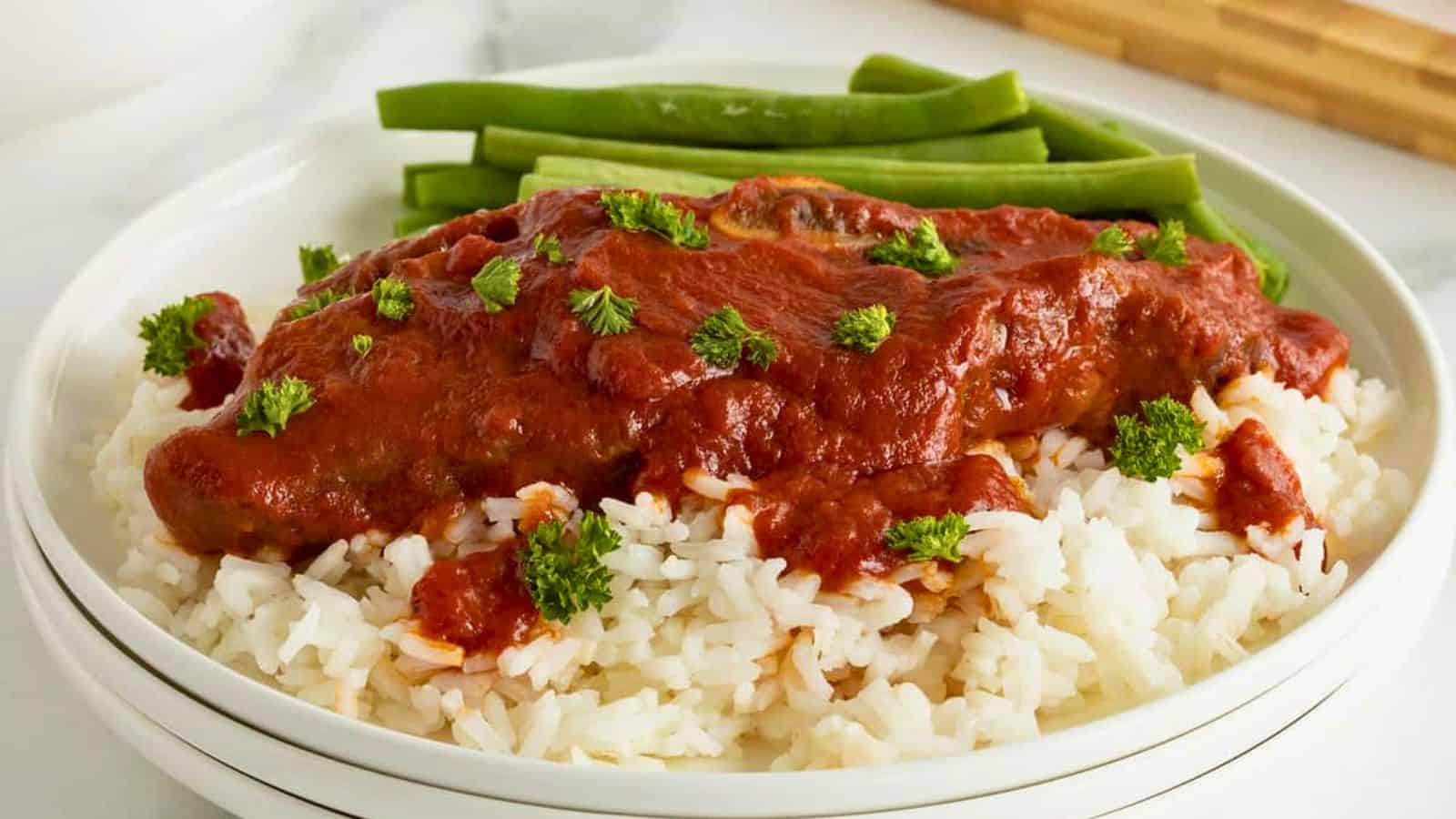 White rice topped with a tomato-based sauce and herbs, served with green beans on a white plate.