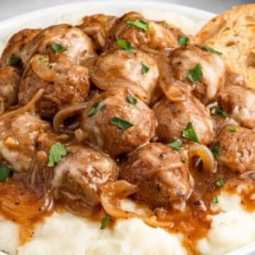 Meatballs in gravy with onions on mashed potatoes, garnished with parsley, served with toasted bread slices.
