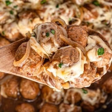 A wooden spoon holds cheesy meatballs with onions and herbs over a pan filled with more meatballs in sauce, perfect for easy Crockpot Meals.