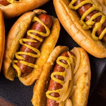 Several hot dogs in buns topped with mustard and onions are arranged on a dark serving platter.