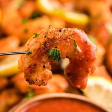 A seasoned, cooked shrimp on a fork with lemon slices and sauce in the background.