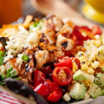 Colorful salad bowl with grilled chicken, avocado, tomatoes, corn, shredded cheese, and greens on a table.