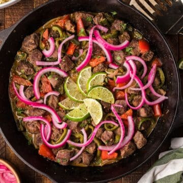A skillet with cooked beef, red onions, peppers, and lime slices, on a wooden table with utensils nearby.