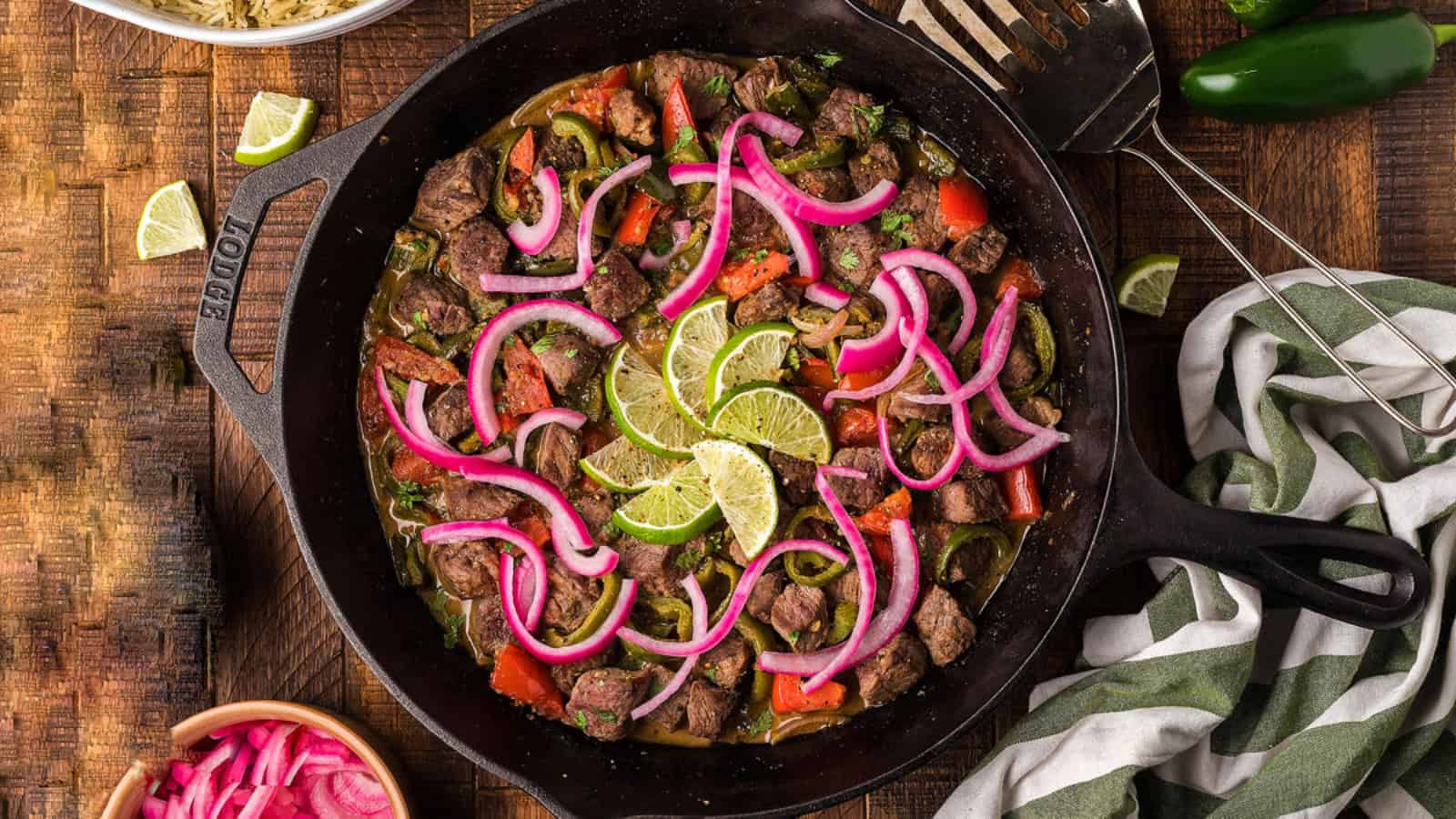 A skillet with cooked beef, red onions, peppers, and lime slices, on a wooden table with utensils nearby.