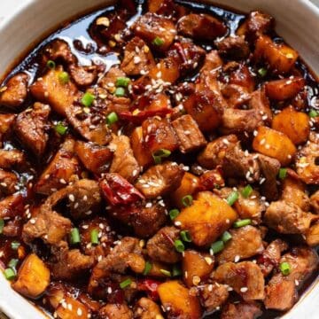 A bowl of braised pork with chunks of pineapple, garnished with sesame seeds and chopped green onions.