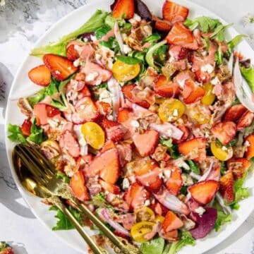 Fresh salad with strawberries, cherry tomatoes, feta, and walnuts on a white plate with strawberry dressing nearby.